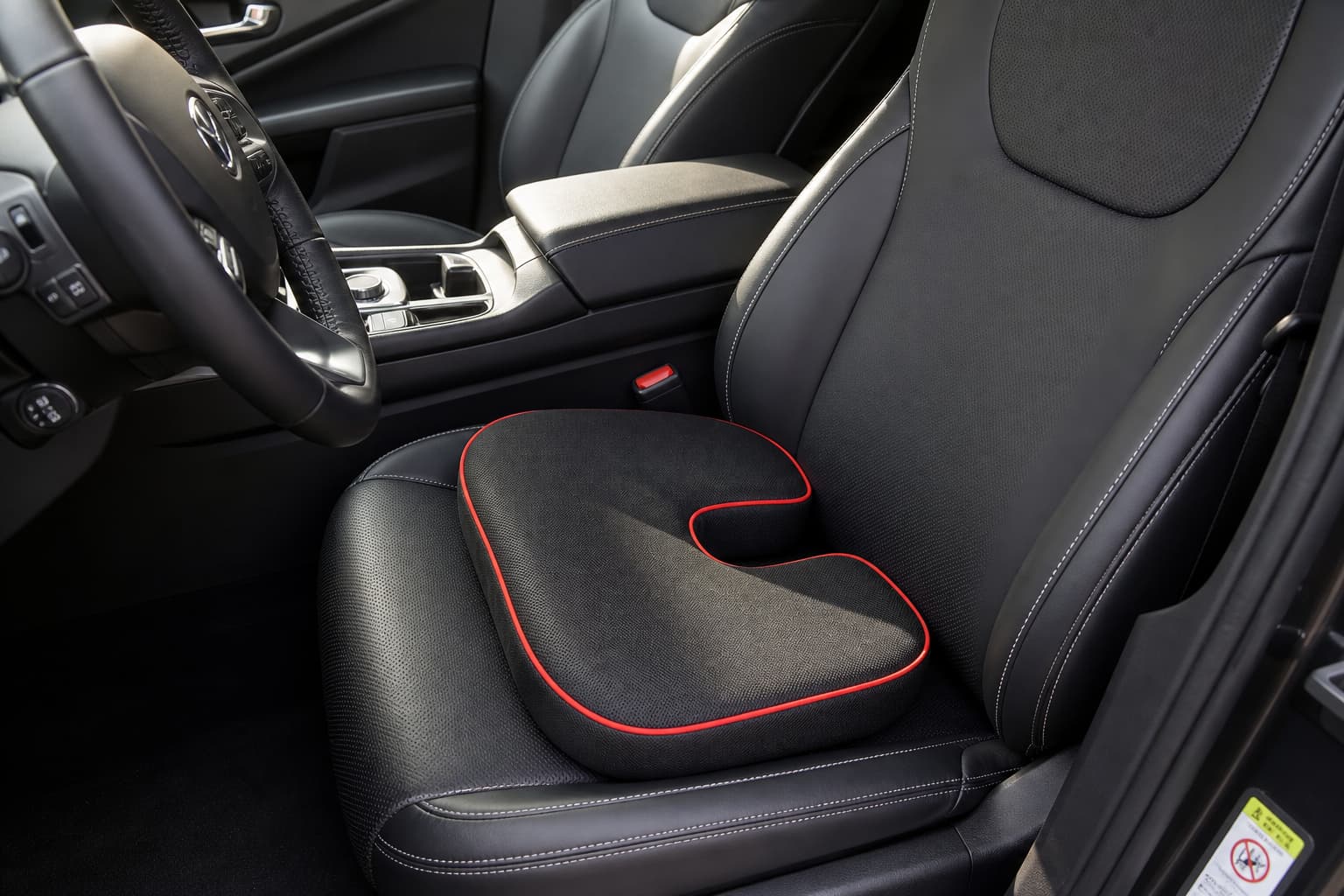 Coussin orthopédique en forme de U, noir avec bordure rouge, posé sur un siège de voiture, lumière naturelle entrant par la fenêtre.